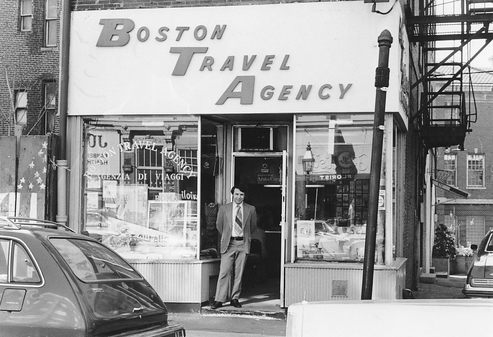 Black and white photo of the original Boston Travel Agency storefront in the North End, with a man in a suit standing in the doorway under the sign